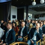 Uczestnicy konferencji biznesowej siedzący w rzędach i słuchający prelegenta podczas wydarzenia w nowoczesnej sali konferencyjnej.