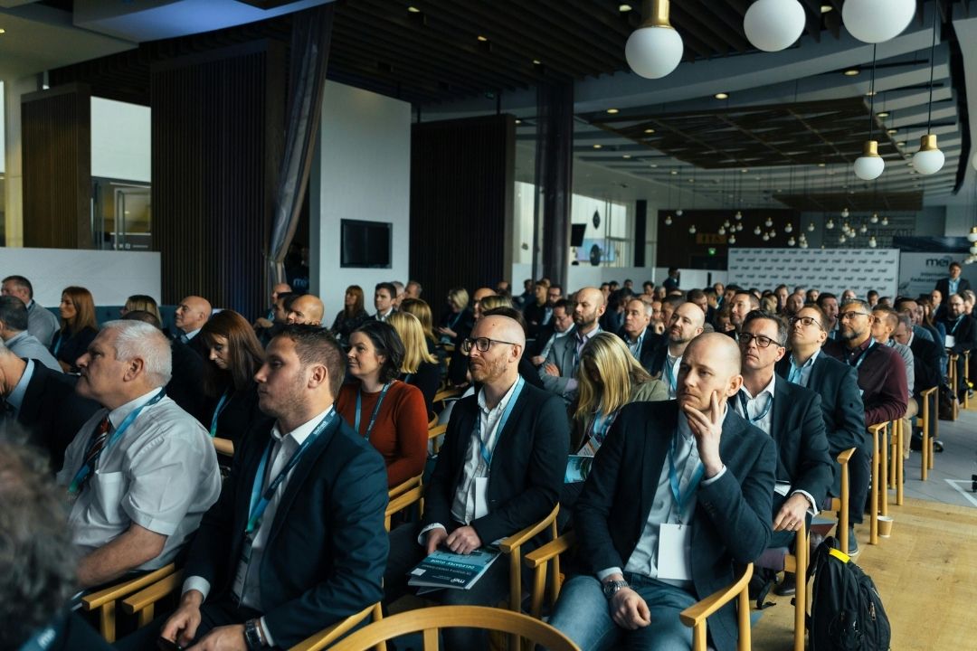 Uczestnicy konferencji biznesowej siedzący w rzędach i słuchający prelegenta podczas wydarzenia w nowoczesnej sali konferencyjnej.